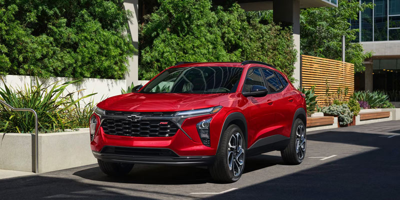 A bright red Chevrolet Trax parked in a modern, landscaped parking area with green plants and urban architecture.