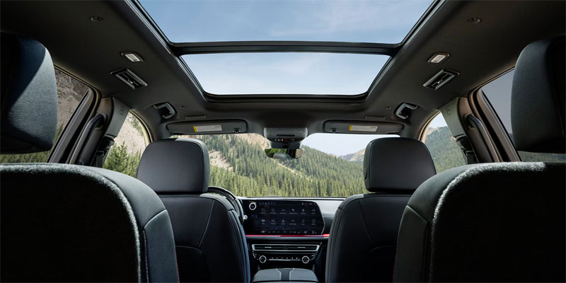 Interior view looking forward from the back seat, highlighting the dark leather upholstery, digital dashboard, and a large panoramic sunroof showing blue sky and green hills.