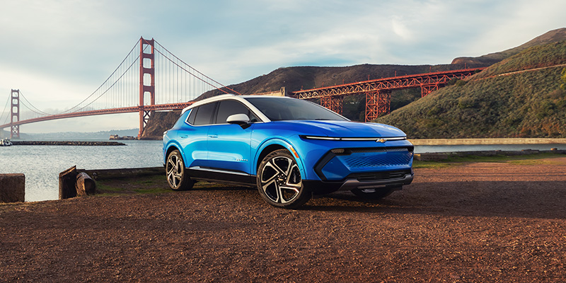 Front-three-quarter view of a bright blue Chevrolet Equinox EV with a white roof parked on a gravel area near the water, with the Golden Gate Bridge visible in the background.